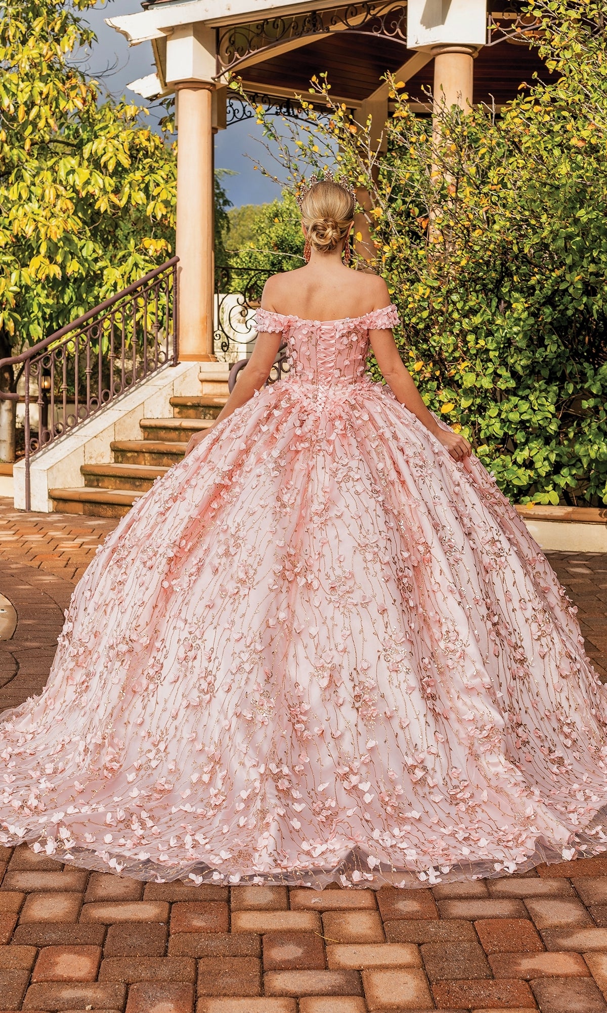 A woman in the Quinceanera Dress 1734 By Dancing Queen, a pink off-the-shoulder ball gown with floral details, stands outdoors on a stone path facing away from the camera.